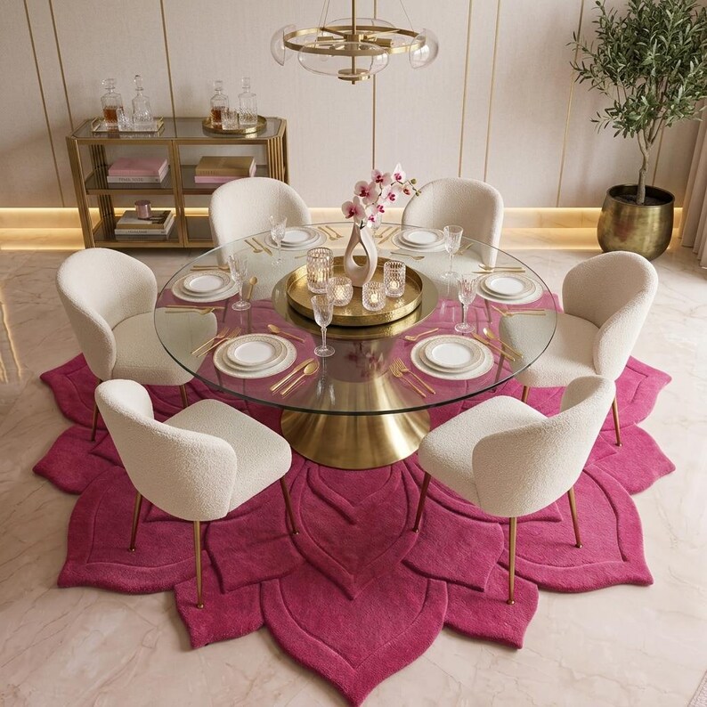 May include: A round glass dining table with a gold pedestal base, surrounded by six cream-colored upholstered chairs. The table is set with white plates, gold cutlery, and a gold tray holding candles. A large, fuchsia, petal-shaped area rug.