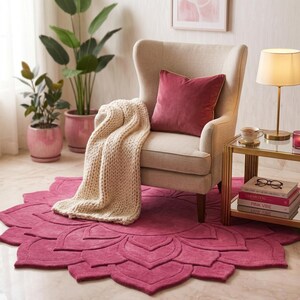 May include: A circular, deep pink rug with a layered petal design, featuring a beige wingback chair, a cream-colored chunky knit throw blanket, a solid dark pink square pillow, and a gold-toned side table with books and a lamp.
