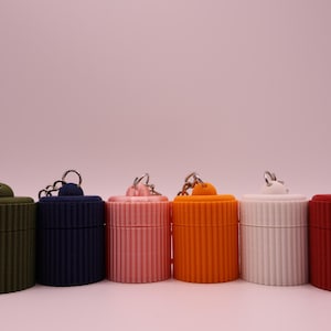 May include: Six small, cylindrical containers in olive green, navy blue, pink, orange, white, and red. Each has a ribbed texture and a silver keychain ring. The background is a pale pink. The containers are likely for small items.