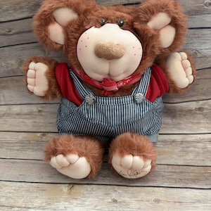 Puede incluir: Un oso de peluche marrón con una camisa roja, un peto a rayas azules y blancas y un pañuelo rojo. El oso tiene una nariz marrón claro, mejillas rosadas con pecas y orejas grandes marrones. Los pies del oso son de color marrón claro con una firma.