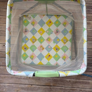 May include: A vintage baby bassinet with a white mesh net and a colorful patterned interior. The bassinet has a light green frame and handles, and the interior features a diamond pattern with cartoon characters in pastel colors.
