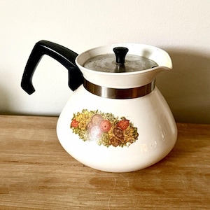 May include: White ceramic coffee pot with a black handle and lid. The pot features a colorful fruit and floral design around the middle, with a silver band. The pot is sitting on a wooden surface.