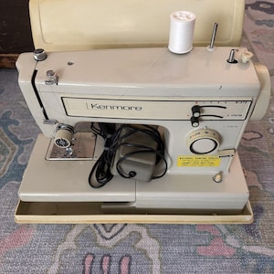May include: A vintage cream-coloured Kenmore sewing machine with a white thread spool. The machine features a built-in light, dials, and the Kenmore logo. It sits on a light cream base and has a black power cord. The machine is a classic sewing tool.