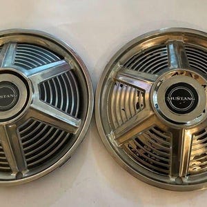 Puede incluir: Dos tapacubos Mustang vintage. Cada tapacubo es plateado con un diseño de estrella y una tapa central negra que dice "MUSTANG". Los tapacubos tienen un patrón circular acanalado alrededor del centro.