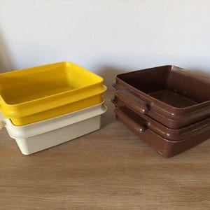 Puede incluir: Una pila de tres recipientes rectangulares de plástico amarillo y blanquecino con asas, junto a una pila de cuatro recipientes marrones. Los recipientes están sobre una superficie de madera.