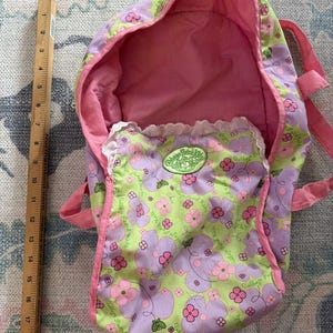 May include: A pink and green floral patterned doll carrier with a hood. The carrier has pink straps and a pink interior. The front pocket has a white lace trim and a green circular logo that says "Cabbage Patch Kids".