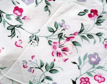 May include: Close-up of a white fabric with a floral pattern. The design features pink and purple flowers with green leaves and stems. The fabric is folded, showing the pattern's continuity.