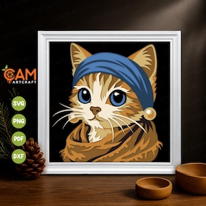 May include: A framed digital art print of a cat wearing a blue cap. The cat has large blue eyes, brown and tan fur, and long white whiskers. The print is in a white frame and sits on a wooden surface. The image includes the text "CAM Artcraft" and file type icons.