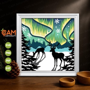 May include: A framed paper cut artwork showing a winter scene with two deer in silhouette. The layered design features a snowy landscape, evergreen trees, and the aurora borealis in green and yellow. The frame is white. The artwork is in a white frame.