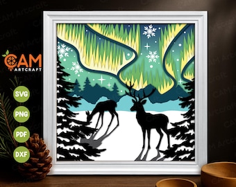May include: A framed paper cut art piece depicting a winter scene with two deer in silhouette. The artwork features a layered design with a snowy landscape, evergreen trees, and the aurora borealis in shades of green and yellow. The frame is white.