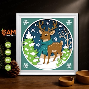 May include: A framed paper cut art piece depicting a deer wearing a teal scarf and festive lights on its antlers, set in a snowy winter landscape. The artwork includes green trees and snowflakes, with the text "CAM Artcraft" and file type icons.