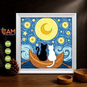 Op de afbeelding: Een ingelijst papieren kunstwerk met een zwart-wit kattenpaar in een boot onder een sterrenhemel met een halve maan. Het kunstwerk zit in een witte lijst en bevat de tekst "CAM Artcraft".