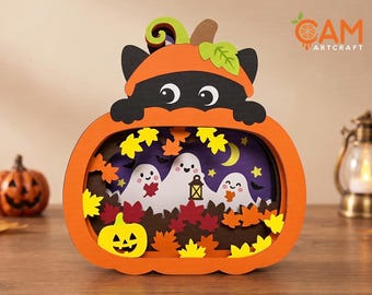 May include: A layered wooden Halloween decoration shaped like a pumpkin with a black cat peeking over the top. The interior features three white ghosts, a yellow crescent moon, stars, bats, and colorful autumn leaves. The text CAM ARTCRAFT is visible.