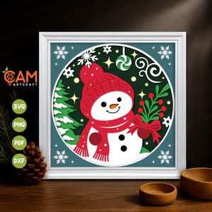 Pode incluir: Uma obra de arte de Natal em papel recortado e emoldurada. A obra apresenta um boneco de neve com chapéu e cachecol vermelhos, segurando um ramo de bagas. O fundo é verde escuro com flocos de neve e uma bengala doce. A moldura é branca.