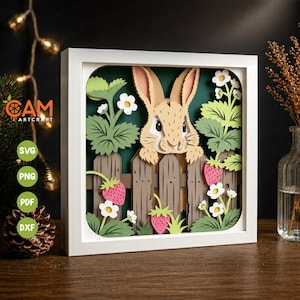 May include: A framed paper art piece depicting a rabbit looking over a wooden fence. The scene includes green leaves, white flowers, and pink strawberries. The artwork is set against a dark green background within a white frame.