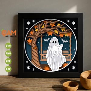 May include: A framed paper art piece. It depicts a white ghost reading a book on a swing beneath a tree with autumnal leaves. Bats are in the background. The artwork is set within a dark blue circle, framed by a black square.