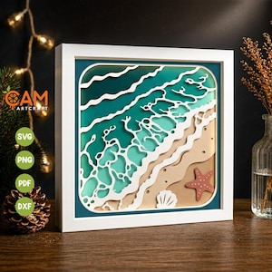 May include: A framed paper art piece depicting a beach scene. The artwork features layers of teal and white waves, a sandy shore, a starfish, and a seashell. The frame is white, and the artwork is displayed on a wooden surface.