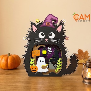Puede incluir: Una decoración de madera recortada en capas con forma de gato negro que lleva un sombrero de bruja morado. El cuerpo del gato tiene un centro hueco que muestra una escena de Halloween con un fantasma blanco, una calabaza y el texto BOO.