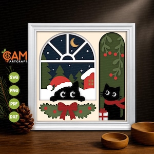 May include: A square white frame containing a layered paper art scene of two black cats. One cat wears a red Santa hat in a snowy window with a crescent moon. The other cat wears a red scarf next to a gift box and green mistletoe branches.