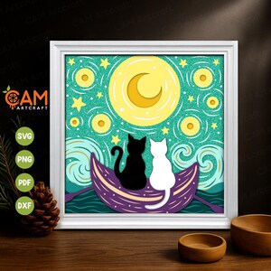 Op de afbeelding: Een ingelijst papieren kunstwerk met twee katten in een boot onder een halve maan en sterren. Het kunstwerk toont een zwarte kat en een witte kat, met een teal glitter achtergrond en een paarse boot. De lijst is wit.