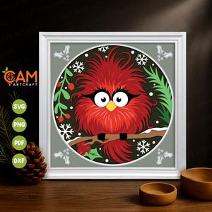 May include: A framed digital art print featuring a stylized red bird with large eyes, perched on a branch. The artwork is set against a dark green background with snowflakes and holly. The frame is white, and the image includes the CAM Artcraft logo.