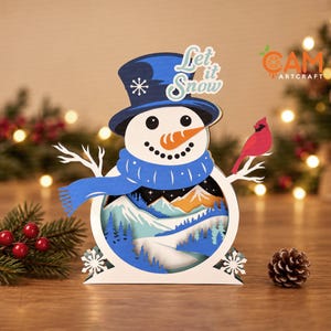 Op de afbeelding: Een gelaagde houten sneeuwpopdecoratie met een blauwe hoge hoed met een witte sneeuwvlok, een blauwe sjaal en een oranje wortelneus. De buik bevat een uitgesneden winterlandschap met bergen en een rode kardinaalvogel op een tak.