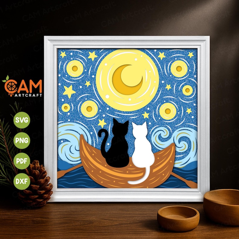 Op de afbeelding: Een ingelijst papieren kunstwerk met een zwart-wit kattenpaar in een boot onder een sterrenhemel met een halve maan. Het kunstwerk zit in een witte lijst en bevat de tekst "CAM Artcraft".