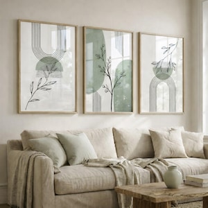 May include: Three framed abstract art prints. Each print features a minimalist design with botanical line drawings, geometric shapes, and a soft green color palette. The artwork is displayed above a beige sofa.