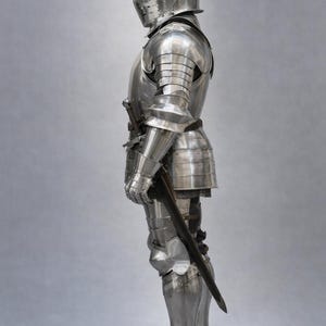 May include: A full suit of polished steel armor, including a helmet with a visor, chest and back plates, arm and leg protection, and gauntlets. A sword is held at the side. The logo "MEDIVAL KNIGHT" is in the upper left corner.