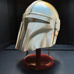 May include: A silver-colored Mandalorian helmet with a dark visor, displayed on a red wooden stand. The helmet has a sleek, metallic finish and is designed with angular features. The stand is round and has a dark base.