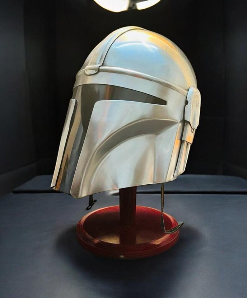 May include: A silver-colored Mandalorian helmet with a dark visor, displayed on a red wooden stand. The helmet has a sleek, metallic finish and is designed with angular features. The stand is round and has a dark base.