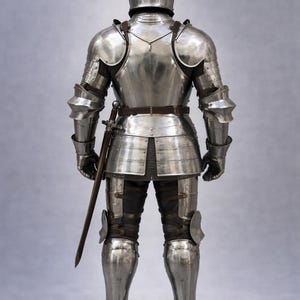 May include: Full suit of polished steel armor, including a helmet, shoulder guards, chest and back plates, and leg protection. A sword is sheathed at the waist. The armor is designed for protection in medieval combat.