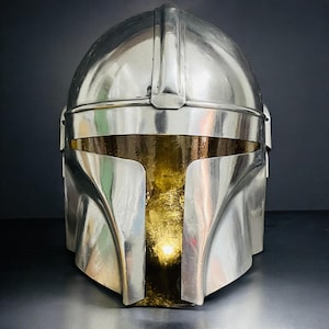 May include: A polished silver helmet with a gold interior, featuring a distinctive T-shaped visor. The helmet's design includes a central ridge and curved cheek plates, reflecting light. The overall aesthetic suggests a futuristic or sci-fi theme.