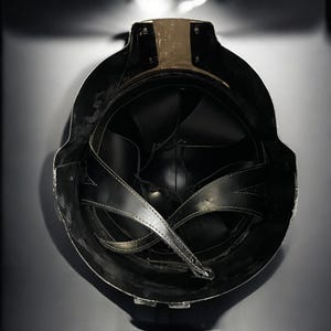 May include: A black helmet with a gold-colored band at the top. The interior features black leather straps and padding. The helmet is designed with a rounded shape and a dark, matte finish.