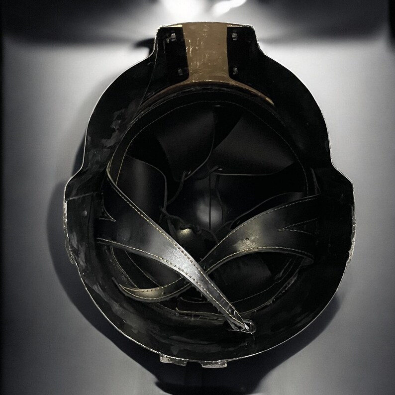 May include: A black helmet with a gold-colored band at the top. The interior features black leather straps and padding. The helmet is designed with a rounded shape and a dark, matte finish.