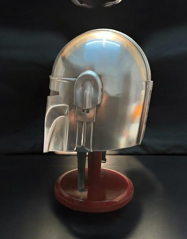 May include: A silver-colored Mandalorian helmet, displayed on a red and brown stand. The helmet has a smooth, metallic finish with detailed features, including a visor and side panels. The stand is round with a dark base.
