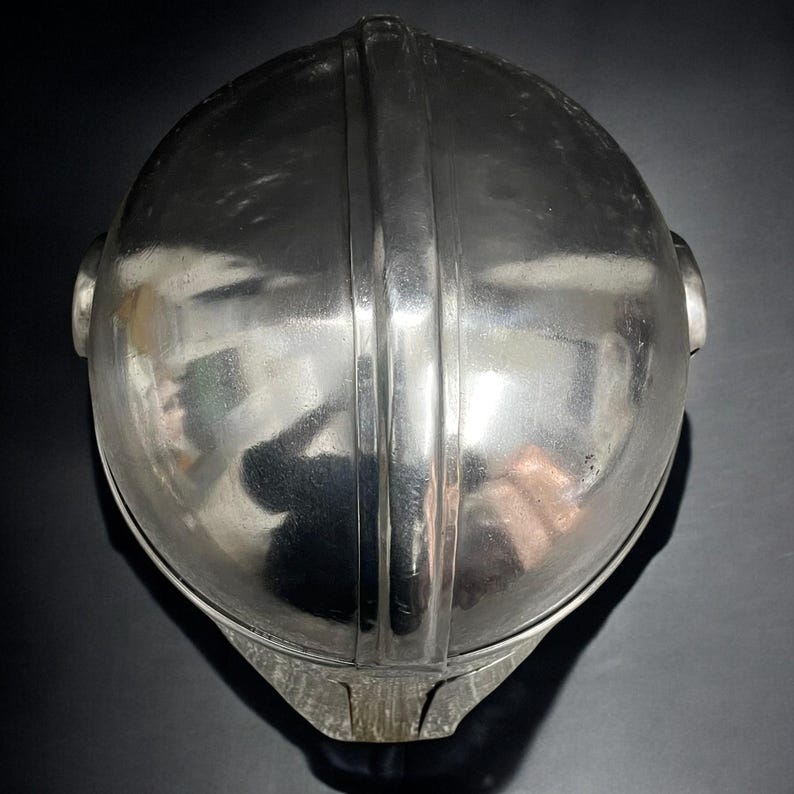 May include: A shiny, silver-colored metal helmet with a rounded top and a vertical ridge down the center. The helmet has a smooth, reflective surface and two small, circular protrusions on the sides. The bottom portion has a textured, matte finish.