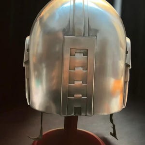 May include: A silver-colored helmet with a rounded top and rectangular features. The back of the helmet has a series of rectangular cutouts. The helmet is displayed on a red stand.