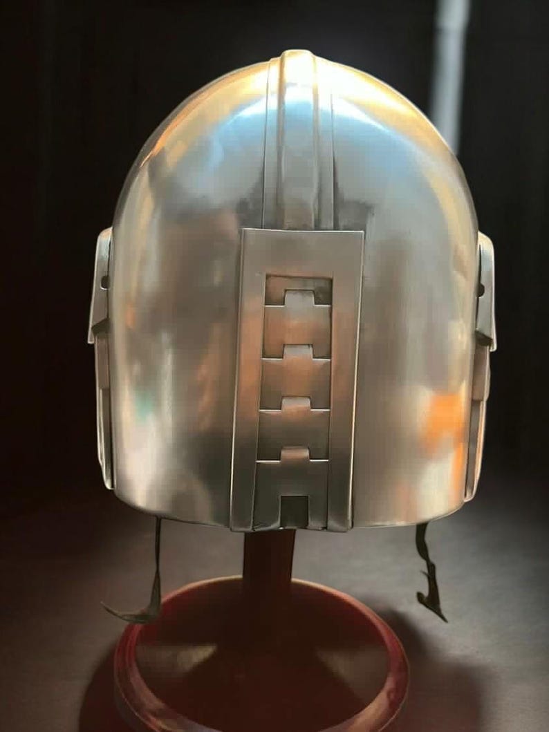 May include: A silver-colored helmet with a rounded top and rectangular features. The back of the helmet has a series of rectangular cutouts. The helmet is displayed on a red stand.