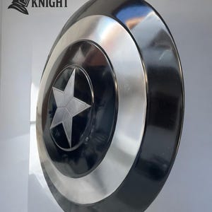 May include: A round shield with a black and silver design. The shield features a central silver star on a black background, surrounded by concentric silver and black bands. The shield has a glossy finish and is likely made of metal.