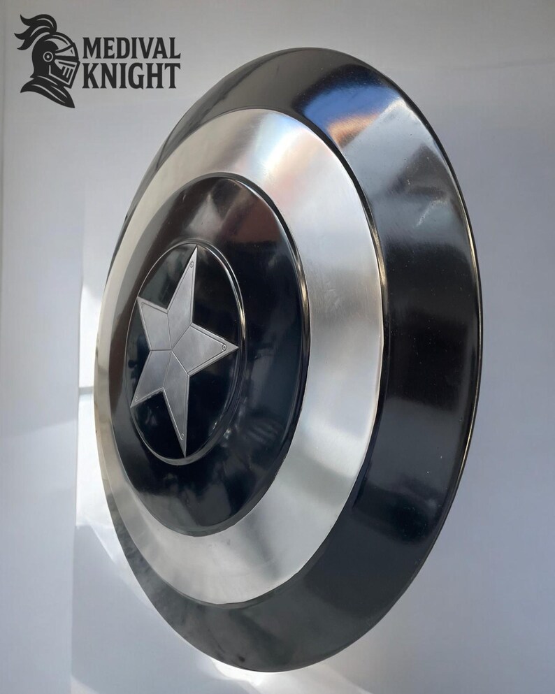 May include: A round shield with a black and silver design. The shield features a central silver star on a black background, surrounded by concentric silver and black bands. The shield has a glossy finish and is likely made of metal.