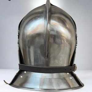 May include: A polished steel medieval helmet with a prominent central ridge and a leather chin strap. The helmet features a rounded crown and a flared lower edge, with rivets securing the components. The "MEDIVAL KNIGHT" logo is visible in the upper left corner.