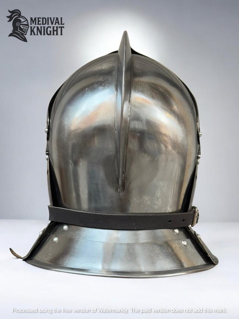 May include: A polished steel medieval helmet with a prominent central ridge and a leather chin strap. The helmet features a rounded crown and a flared lower edge, with rivets securing the components. The "MEDIVAL KNIGHT" logo is visible in the upper left corner.