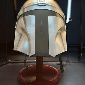 May include: A silver-colored Mandalorian helmet with a dark visor, displayed on a red stand. The helmet has a distinctive T-shaped visor and is made of a metallic material. The stand is round and has a dark red finish.