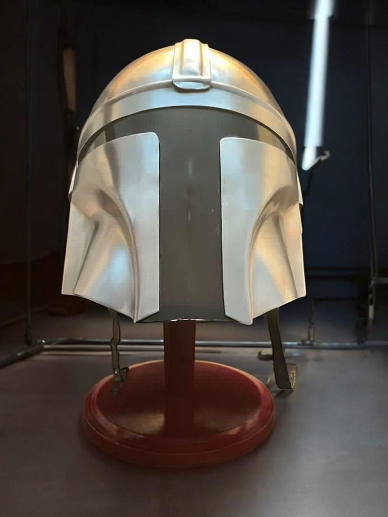 May include: A silver-colored Mandalorian helmet with a dark visor, displayed on a red stand. The helmet has a distinctive T-shaped visor and is made of a metallic material. The stand is round and has a dark red finish.