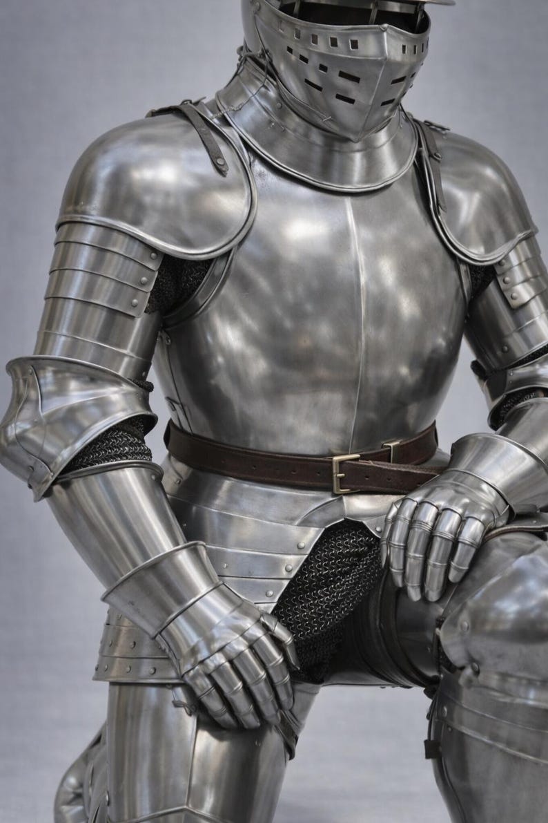 May include: A detailed shot of a medieval suit of armor. The polished steel armor includes a helmet, chest plate, shoulder guards, arm and leg protection, and gauntlets. A brown leather belt is fastened around the waist. The armor is displayed against a neutral background.