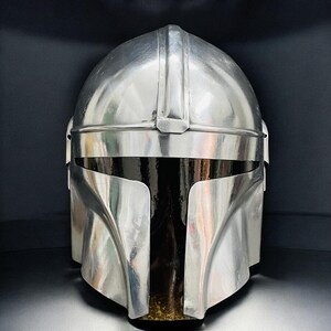 May include: A metallic silver Mandalorian helmet with a sleek, polished finish. The helmet features a distinctive T-shaped visor and a rounded top, reflecting the surrounding environment. The design is reminiscent of science fiction and space opera themes.