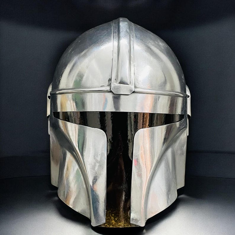 May include: A metallic silver Mandalorian helmet with a sleek, polished finish. The helmet features a distinctive T-shaped visor and a rounded top, reflecting the surrounding environment. The design is reminiscent of science fiction and space opera themes.