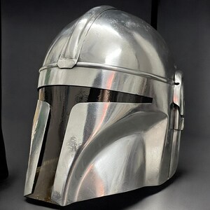 May include: A metallic silver helmet with a dark visor. The helmet has a smooth, reflective surface with visible seams and a rounded top. The design features a prominent brow ridge and cheek plates, suggesting a futuristic or sci-fi theme.