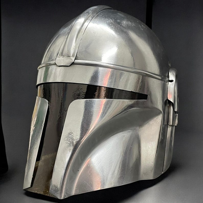 May include: A metallic silver helmet with a dark visor. The helmet has a smooth, reflective surface with visible seams and a rounded top. The design features a prominent brow ridge and cheek plates, suggesting a futuristic or sci-fi theme.